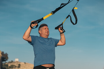Frontal image of an older overweight white-haired person performing suspension training on the...
