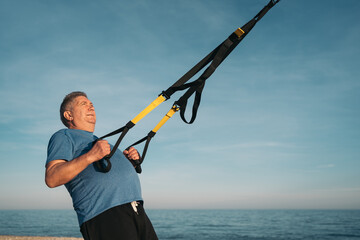 Side image of an older overweight white-haired person performing suspension training on the beach carrying her arms backwards to strengthen her back with the exercises.