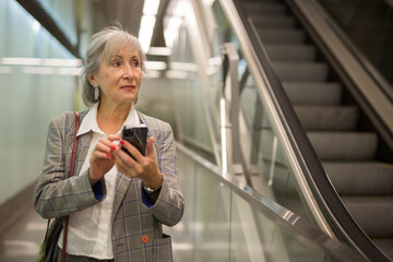 Mature businesswoman walking in office building beside escalator and using her smartphone