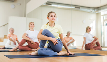 Teenage girl sitting in lord of fisher pose during group yoga training in studio.