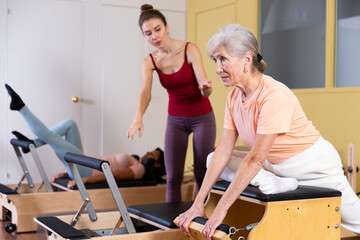 Fototapeta na wymiar Mature European woman practicing Pilates in a group workout with a female instructor trains on a combined chair in a ..fitness studio