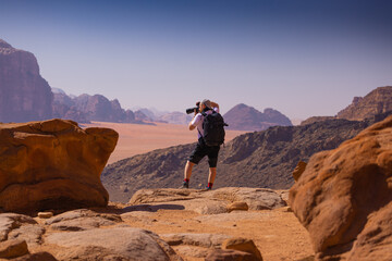 Wadi Rum w Jordanii. Mężczyzna stojący na skale fotografujący pustynię. - obrazy, fototapety, plakaty