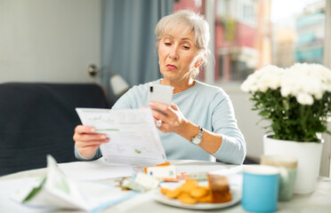 Focused senior woman reading documents at home table and using cell phone to make copy of papers.