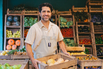 Middle-aged Latin man with a smile holding a crate of squash in a greengrocer's shop while looking at the camera. - Powered by Adobe