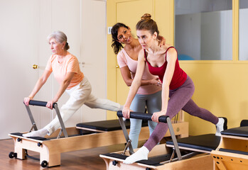 Fototapeta na wymiar Two active women practicing Pilates in a fitness studio perform an exercise using a reformer bed, where a female instructor ..helps them do it correctly