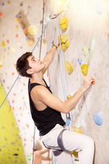 Fototapeta premium young muscular guy, insured by trainer, ascend up artificial climbing wall indoors. rappelling athlete