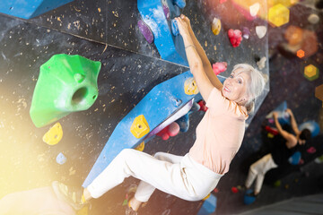 Positive focused mature woman exercising on black climbing wall without safety belts and preparing...