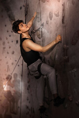 Fototapeta na wymiar Positive extreme young guy with rope training on concrete climbing rock wall to maintain healthy discipline in indoor adventure park
