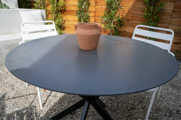 Dark circular patio table with clay pot on top and white metal chairs on a gravel lined patio.