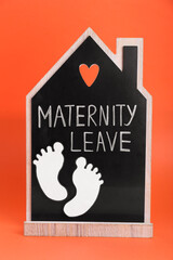 Wooden house figure with words Maternity Leave and paper cutout of baby feet on orange background