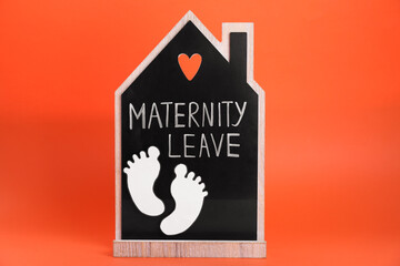 Wooden house figure with words Maternity Leave and paper cutout of baby feet on orange background