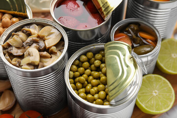 Open tin cans with different preserved products on wooden board, closeup