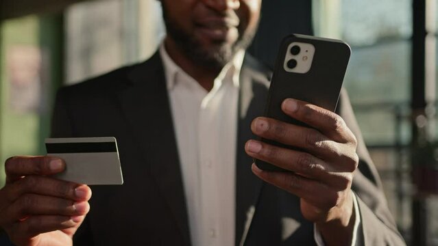 Close up credit bank card unknown African American adult man businessman banker in office pay online on mobile phone money finances paying shopping ecommerce enter secure code safe transaction payment