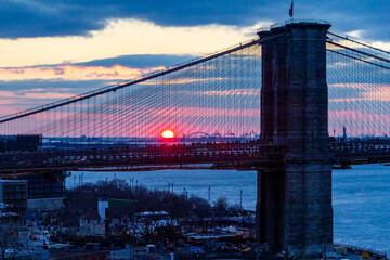 Sunset on the horizon behind the Brooklyn Bridge with crowds of people gathered to watch the setting sun in New York City
