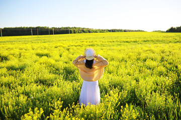 Pretty girl with a hat in her hand walks in a field with field flowers and smiles sincerely