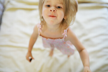 A small child with blond hair lies on the bed. Baby girl is playing on the couch.
