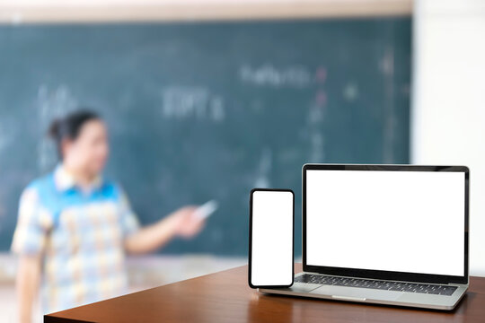 front view Blank screen of black digital laptop and smartphone on wooden table, teacher standing in front of classroom. educational software website technology