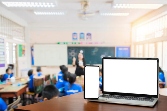Front view, blank screen of black digital laptop and smartphone with tablet on the wood table. Children students in classroom background. Education software website technology ads concept.