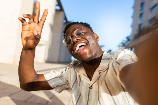 African american man with glasses taking selfie outdoors looking at camera waving hand.