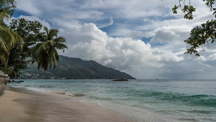 The waves of the turquoise ocean are foaming on the beach. Wet sand glistens. Green branches and palm trees against a  blue sky and clouds. A hill in the distance. Seychelles. Mahe. Beau Vallon