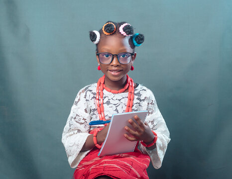 African Nigerian female girl child or student with eye glasses, wearing yoruba local dress attire and beads, holding a pen and note book while smiling and looking at the camera