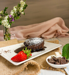chocolate coulant, strawberries and mint leaves on a white plate on a wooden table with flowers and a pink silk cloth.