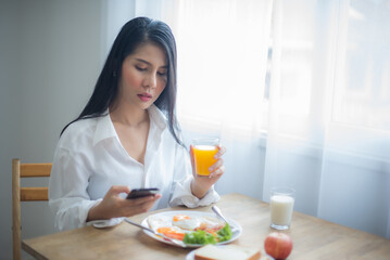 Obraz na płótnie Canvas Beautiful asian woman eyes look on the screen through emails and messages on her phone as she picked up a glass of juice and breakfast on the table.