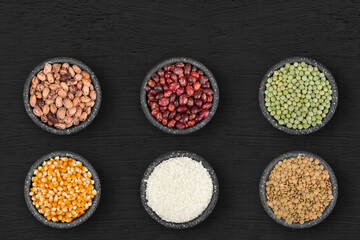 Set of bowls with various grains, lentils, corn, rice, beans and peas, on a black wood.
