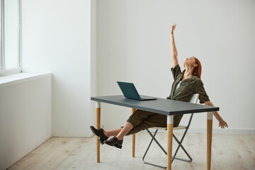a very happy woman sits at a laptop and rejoices at the completed tasks raising her hands up