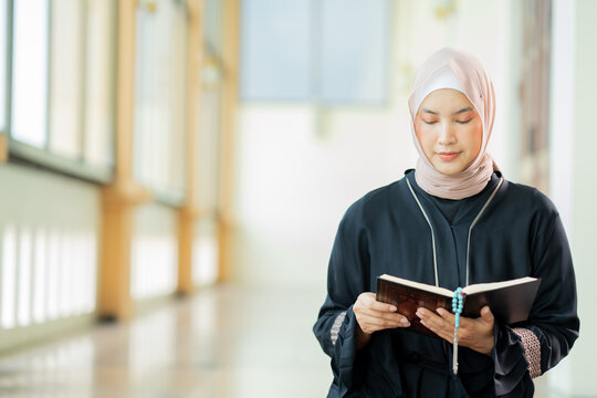 The image of an Asian Muslim woman in the Islamic religion in hijab in cream and black color. reading the Quran in a beautiful mosque, Arabic word translation: The Holy Al Quran (holy book of Muslims)