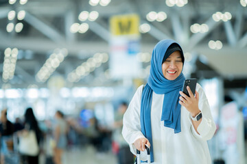 Fototapeta na wymiar An Asian Muslim wearing a blue hijab is preparing for a vacation and she is at the airport. She is using her mobile phone to contact her friends and Muslim travelers.