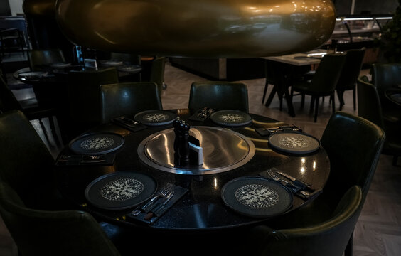 BATUMI, GEORGIA - MARCH 11, 2023: Bakir Home Restaurant black wooden round table served and dark chairs around it. the dome of a metal brass air extractor hangs over the table. dusk new interior.