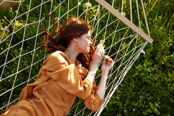 a beautiful, elegant woman lies in a long orange dress on a mesh hammock resting in nature,...
