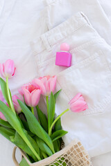 Pink tulips in macrame bag and perfume bottle on white jeans background