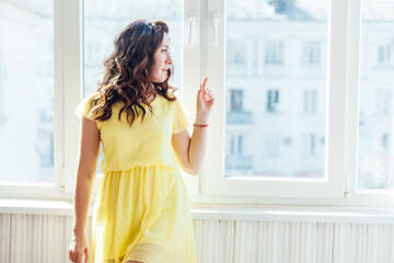 beautiful brunette woman in a yellow dress stands at a large window