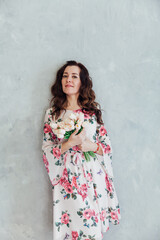 Fototapeta premium a brunette woman in a floral dress stands with flowers against a grey wall