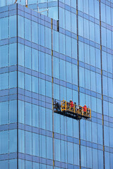 Builders in red overalls are mounting the glass facade of a multi-storey building on a suspended cradle. - 593457213