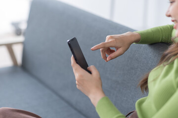Young student asian woman wearing headphones holding cell phone while lying on sofa in bright apartment