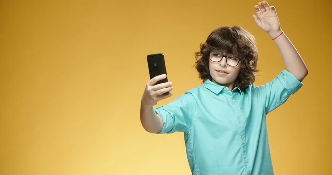 Cute little boy using his mobile phone to take pictures or selfies and positively smiling, isolated on yellow background - always online, emotions concept close up