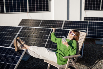 Young woman talks on phone while sitting relaxed on rooftop with a solar station. Happy owner of...