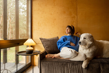 Portrait of a young woman with her dog sitting together on a couch and looking at window with view...