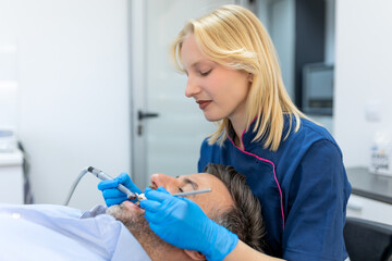 Dentist in blue uniform doing teeth procedure to patient, male patient during treatment procedure in contemporary dental clinic