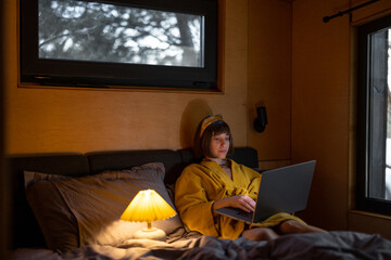 Woman works on laptop while lying in bed late in the evening at tiny bedroom of wooden cabin on nature. Online work from cozy house during bedtime