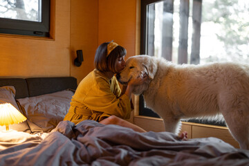 Young woman cares her huge adorable white dog while lying in bed in tiny bedroom of wooden cabin on...