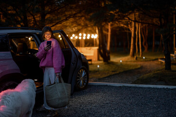 Woman arrives by car to a house in forest, standing with bag and phone near vehicle in the evening...