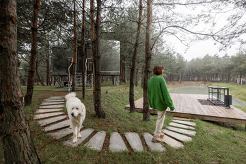 Woman walks on wavy pathway near lake and invisible cabin in pine forest. Connection with nature and sustainability concept. Rest in tiny cabins on nature