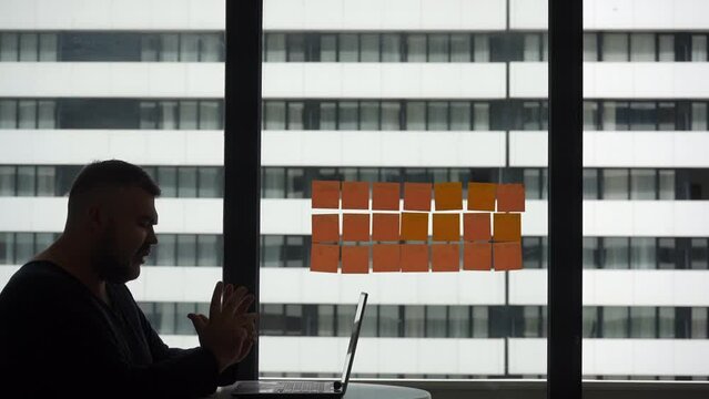 Silhouette of middle aged man with a laptop sitting near a panoramic window on a terrace balcony at home. A man gesticulates with his hands while communicating via video call. Video conference online.
