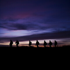 Soldiers walking at dusk silhouette, their forms fading into the darkening blues and purples of the sky. The peaceful serenity of the evening is juxtaposed with the gravity of their situation.
