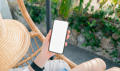 Woman sitting on the chair near swimming pool with hand holding smartphone mockup of blank screen,...