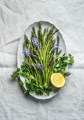 Delicious raw green asparagus on plate with springtime flowers, lemon and herbs, top view. Spring...